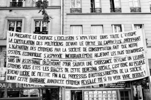 A large banner with French text is hung between buildings in a city street. The banner discusses topics like economic struggles, political critiques, and societal issues, addressing the impact of capitalism, unemployment, exclusion, and the erosion of democracy. It also references the Manifestation on 16 septembre 2010 against réforme des retraites.