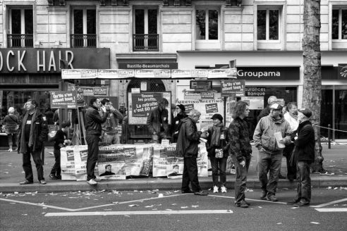 A black-and-white photo of a group of people gathered in front of a newsstand, with signs and posters related to the réforme des retraites. Some individuals are conversing, reflecting on the manifestation that took place on 16 septembre 2010, while others stand looking on. Storefronts and a tree-lined sidewalk are visible in the background.