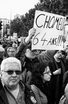 A black-and-white photo showing a manifestation with numerous participants. A prominently visible demonstrator holds up a sign reading "CHOMEUR 64 ans!!!" Other protesters, some with glasses, appear focused and engaged in the background. The atmosphere on 16 septembre 2010 seems serious and determined.