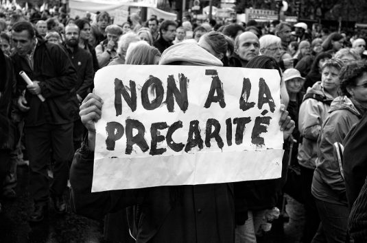A person holds a sign that reads "Non à la Precarité" (No to Precarity) in French, amidst a densely packed crowd of protesters during the 16 septembre 2010 manifestation. The black-and-white image captures the determination of the demonstrators rallying against the réforme des retraites.