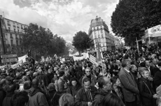 A black and white photo captures a large crowd gathered for a manifestation on a city street on 16 septembre 2010. They are holding signs, banners, and flags advocating for a réforme des retraites. Surrounding buildings and trees frame the scene, emphasizing the serious and determined mood.