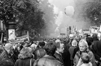 Black-and-white image of a crowded street protest, likely during the manifestation on 16 septembre 2010. People holding signs and banners march in a densely packed area surrounded by trees. Smoke and balloons are visible in the background, reflecting various emotions tied to the réforme des retraites.