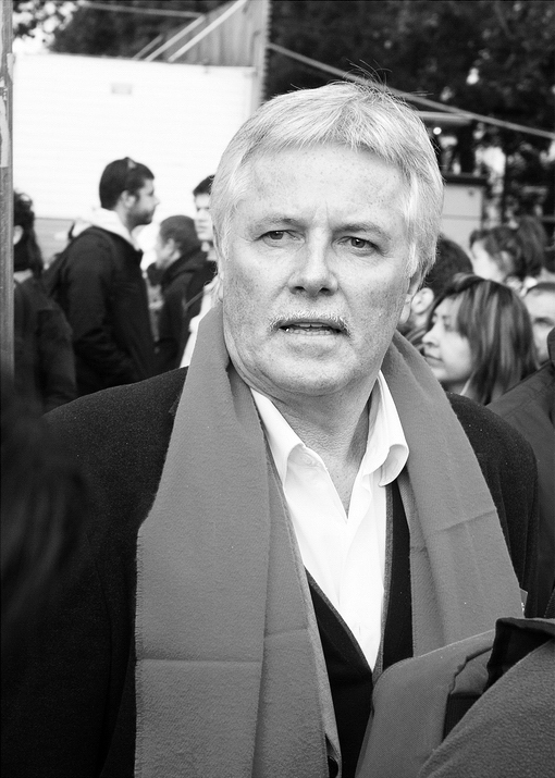 A black and white photo of a person with short, light hair and a mustache, wearing a dark coat and a light-colored scarf. They appear to be speaking or reacting during the manifestation on 16 septembre 2010, against the réforme des retraites, with several other people visible in the blurred background.