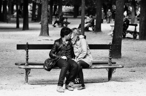A couple sits on a park bench, kissing and embracing, surrounded by trees and people on other benches in the background. In this black and white image, it’s clear that moments like these are toujours mieux à deux.