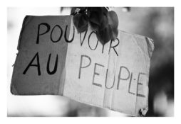 Black and white image of a cardboard sign that reads "POUVOIR AU PEUPLE," with some handwritten letters darker than others. The sign is hanging, and there are leaves partially covering its top portion. This iconic phrase, seen during the marches populaires of 21 Avril 2012, means "Power to the People.