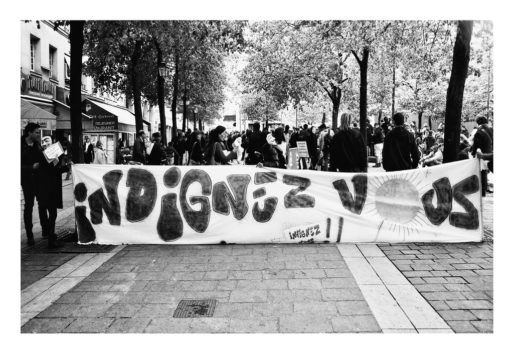 Black and white photo of a public protest. Individuals stand behind a large banner that reads "Indignez Vous" in bold, stylized letters. Engaged in discussion, people hold signs. The scene is set in an outdoor urban area with trees and buildings, capturing the Manifestation on 21 Avril 2012 in soutien aux marches populaires.