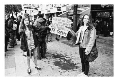 A black-and-white photo of two people holding a protest sign in a busy street during the manifastation on 21 Avril 2012. The sign reads, "Tout ce qu'ils veulent, c'est une France qui ferme sa gueule," meaning "All they want is a France that shuts up." Other protestors and shops are visible in the background.