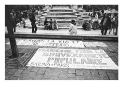 Black and white image of a public square with a group of people sitting around a fountain. Banners on the ground read "MARCHÉ POUR LA SOUVERAINETÉ POPULAIRE" and "PRÉSERVONS LA VIE ET LES BIENS COMMUNS." Trees and a pirate flag are visible in the background, marking the manifestation on 21 Avril 2012.