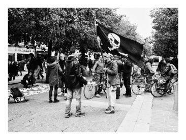 A black and white photo of a group of people gathered outdoors during a manifestation. One person holds a large flag with a skull and crossbones symbol. Some people have bicycles, and others are standing or sitting on the pavement. Trees are in the background along with some buildings.