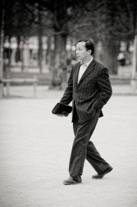 A man in a veste rayée walks outdoors with one hand in his pocket and a briefcase in the other. Leafless trees and park rails are visible in the background, highlighting timeless mode in this black and white image.
