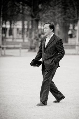 A man in a veste rayée walks outdoors with one hand in his pocket and a briefcase in the other. Leafless trees and park rails are visible in the background, highlighting timeless mode in this black and white image.