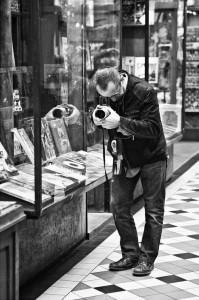 A man in a jacket, possibly a passionné de photographie, leans down with his Hasselblad to photograph books displayed behind glass in a shop window. The black-and-white scene features a checkered tile floor.