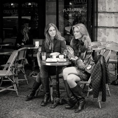 Plat du jour Two women sit at a small café table outdoors, enjoying drinks, pastries, and the plat du jour. Stylishly dressed and deep in conversation, they relax in wicker chairs as other patrons and the café’s name on the window complete the charming scene.