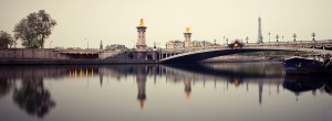 Panoramic view of the Pont Alexandre III bridge in Paris, featuring ornate lampposts and golden statues, with the Eiffel Tower visible in the background. The bridge spans the calm waters of the River Seine, with poses longues capturing reflections of the structures in the water along quais de Seine.