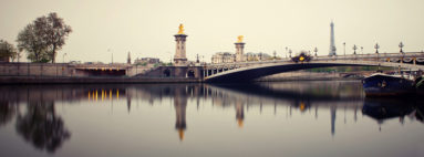 Panoramic view of the Pont Alexandre III bridge in Paris, featuring ornate lampposts and golden statues, with the Eiffel Tower visible in the background. The bridge spans the calm waters of the River Seine, with poses longues capturing reflections of the structures in the water along quais de Seine.