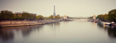 A serene view of the Seine River in Paris. The Eiffel Tower stands tall in the hazy background. The quais de Seine are lined with trees and buildings, and several boats are docked along the right side. A bridge spans the river, decorated with statues, captured beautifully through poses longues photography.