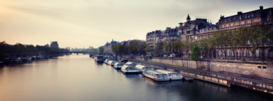 A serene view of the Seine River in Paris during dusk, with riverboats lined up along the quais de Seine. Historic buildings with classic French architecture flank the river. The calm water reflects the soft light of the fading sun, creating a tranquil urban landscape.