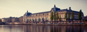 A panoramic view of the Musée d'Orsay in Paris, France, showcasing the historic building along the quais de Seine at dusk. The museum is lit with a warm glow, and a row of trees lines the riverbank in front of the building.