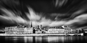 Black-and-white photo of Paris buildings along the Seine River, featuring dramatic clouds and the spire of Notre-Dame Cathedral rising in the center, with reflections visible on the calm water.