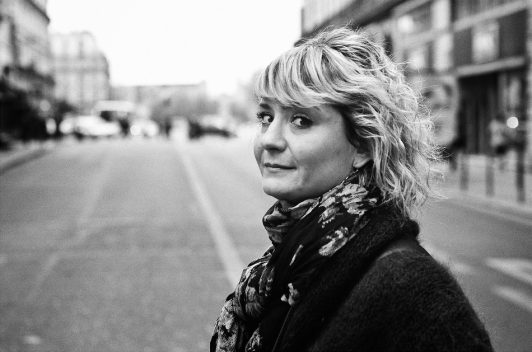 A woman with wavy blonde hair, la femme au foulard, looks over her shoulder at the camera, standing in the middle of a city street on a cloudy day. The image is black and white.