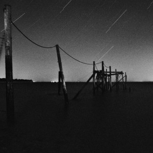 A wooden, weathered vieux carrelet pier extends into dark, calm water beneath the nuit sky, where visible star trails softly illuminate the scene, creating a moody, atmospheric effect.