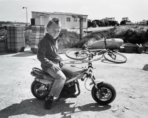 A young child with a styled mohawk hairdo sits on a small, parked minimoto. The scene appears to be an outdoor industrial area with various equipment, a building in the background, and bicycles lying on the ground nearby.