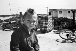 Black and white photo of a young boy with a mohawk hairstyle sitting beside a Minimoto. He is wearing a zip-up jacket. Behind him are stacked lobster traps and another bicycle lying on the ground. In the background, there’s a body of water and a small building.