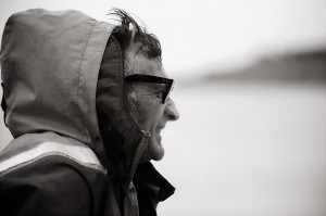 Black-and-white photo of a person in profile, wearing a hooded raincoat and dark sunglasses. The blurred background hints at an overcast or rainy setting, reminiscent of the rugged weather on île d'Aix. The person appears to be outdoors, possibly facing windy or harsh conditions like seasoned marins.