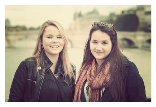 Two women, Cécile and Océane, stand outdoors, smiling at the camera. Cécile has blonde hair and wears a dark jacket, while Océane has long brown hair and sports a scarf with her dark jacket. Behind them, a blurred river scene features a bridge and indistinct buildings.