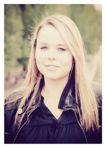 A young woman named Océane, with long, straight blond hair, stands outdoors wearing large hoop earrings and a black leather jacket. She has a serene expression and the background is softly blurred, consisting of green foliage.