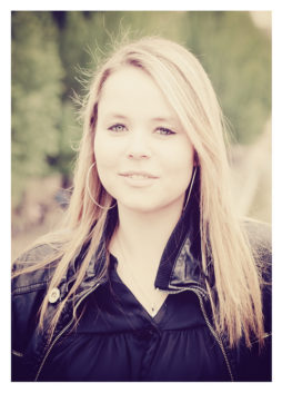 A young woman named Océane, with long, straight blond hair, stands outdoors wearing large hoop earrings and a black leather jacket. She has a serene expression and the background is softly blurred, consisting of green foliage.