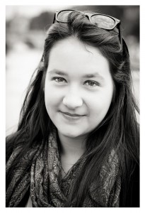 A black-and-white portrait of a person with long, dark hair, smiling slightly. They are wearing a patterned scarf and have sunglasses resting on top of their head. The background is blurred, focusing attention on Cécile's face.