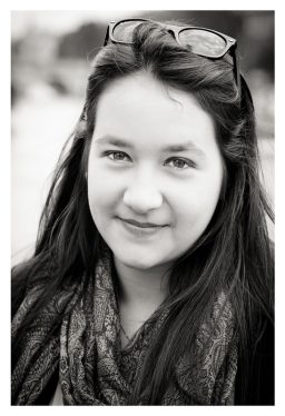A black-and-white portrait of a person with long, dark hair, smiling slightly. They are wearing a patterned scarf and have sunglasses resting on top of their head. The background is blurred, focusing attention on Cécile's face.