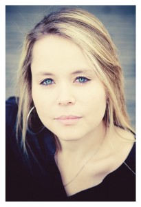 A woman with long, blonde hair and light blue eyes looks directly into the camera. Wearing a black top and silver hoop earrings, Océane exudes elegance. The background is blurred, bringing focus to her face and serene expression.