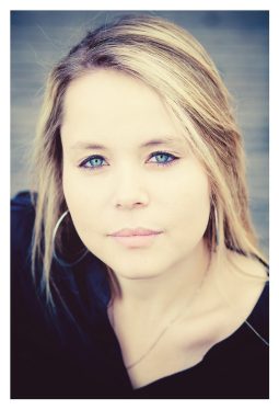 A woman with long, blonde hair and light blue eyes looks directly into the camera. Wearing a black top and silver hoop earrings, Océane exudes elegance. The background is blurred, bringing focus to her face and serene expression.