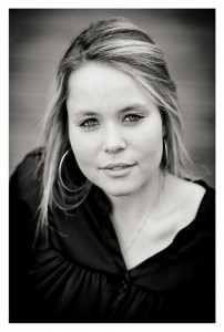 Black and white close-up portrait of a young woman with light hair and medium-length, loose strands. Océane is wearing hoop earrings and a dark, gathered blouse, looking directly at the camera with a calm expression. The background is blurred.