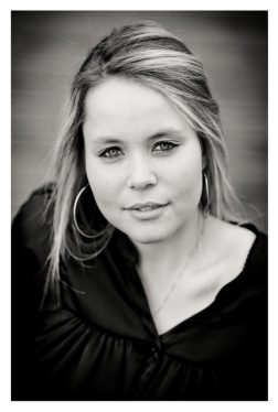 Black and white close-up portrait of a young woman with light hair and medium-length, loose strands. Océane is wearing hoop earrings and a dark, gathered blouse, looking directly at the camera with a calm expression. The background is blurred.