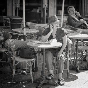 An older woman sits at an outdoor café table, engaged in a phone conversation, with a bag and newspaper in front of her. In the background, a man is also on his phone, seated among empty wicker chairs.