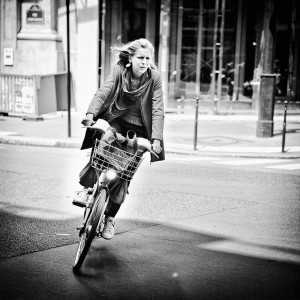 A woman with long hair rides a bicycle with a basket on a city street. Je tourne, she appears focused in her coat and scarf, navigating the black and white scene.