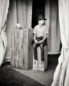 A jeune fille in a top hat, or chapeau, and casual clothing sits on a wooden pedestal next to a taller one adorned with small objects, all set against a curtain backdrop in this black-and-white photo.