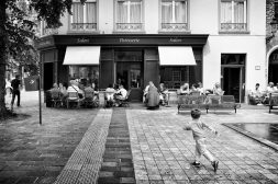 Les enfants courent devant la pâtisserie A black-and-white photo of people sitting at outdoor cafe tables in front of a pâtisserie, while enfants play and a young child runs across the cobblestone square in the foreground.
