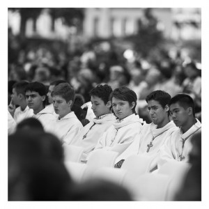 A group of young men, some wearing white robes and cross necklaces, sit closely together outdoors, seemingly engaged in a formal or religious ceremony. The event appears to be ordinations at Notre-Dame with many attendees in the background. The photo is in black and white.