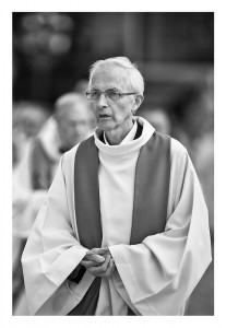 A grayscale image of an elderly person dressed in religious robes at Notre-Dame. They have short, white hair, wear glasses, and appear to be deep in thought. The background is blurred, with other figures faintly visible, emphasizing the solemn and serene atmosphere.
