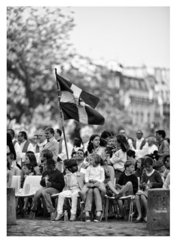 20120630-_MG_0453_MOD_BW_FRAME_resize A group of people, including prêtres, sit on chairs outdoors in a crowded setting, with a large flag held up in the middle featuring bold horizontal and vertical stripes. The background shows trees and buildings. The image is in black and white and evokes the historic ambiance of Notre-Dame during Ordinations.