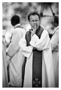 A black and white photo features a priest in religious vestments, including a robe and a stole with a cross emblem. The person has one hand lifted with a finger to their lips, suggesting silence. Other prêtres in similar attire are visible in the background, reminiscent of an ordination ceremony at Notre-Dame.