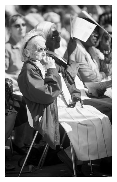 20120630-_MG_0493_MOD_BW_FRAME_resize Black and white photo of an elderly woman sitting in a crowd outdoors, possibly at Notre-Dame during ordinations. She is wearing sunglasses, a headband, and a light-colored coat, holding a piece of cloth. Others around her have various expressions, some with papers over their heads.