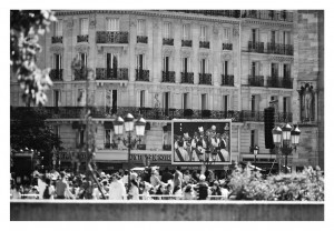 A black and white image shows a bustling crowd in front of an ornate building. The building, reminiscent of Notre-Dame with its intricate balconies and large windows, stands tall. In the background, a large screen displays people in costumes. Street lamps and trees are visible in the foreground.