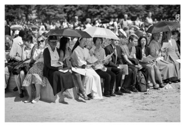 20120630-_MG_0501_MOD_BW_FRAME_resize A black and white photo of a group of people sitting outdoors on a bench on a sunny day. Some individuals, possibly prêtres, hold umbrellas and fans, shading themselves from the sun. They are dressed in a mix of formal and casual attire, with various onlookers visible in the background near Notre-Dame.