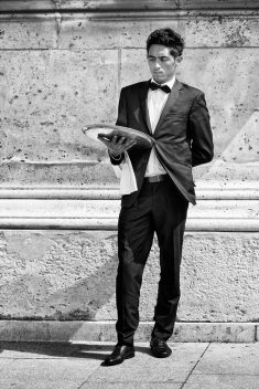 A Garçon Parisien in a formal suit and bow tie stands against a stone wall, holding a serving tray with one hand and a towel draped over his arm, looking serious and slightly pensive.