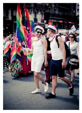 Two men dressed in white tank tops, shorts, and sailor hats walk during a parade. Adorned with colorful leis around their necks and smiling at each other, they embody the name of love. Rainbow flags and a crowd of people are visible in the background, contributing to the festive atmosphere.